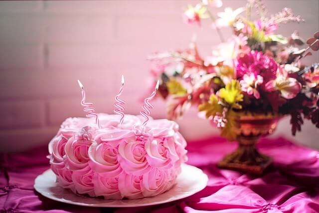 A pink rose-swirled birthday cake with spiral candles on a magenta tablecloth accented by a floral arrangement