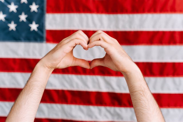 Small business owner in North Texas forming a heart shape with hands in front of an American flag, symbolizing customer appreciation and loyalty.